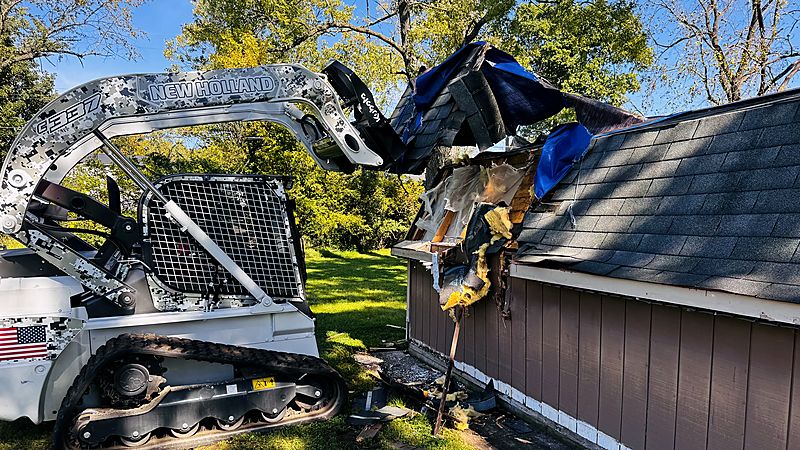 Big Shot Roofing & Restoration New Holland C337 tracked skid steer with digital camo wrap and hydraulic grapple tearing down a damaged roof structure